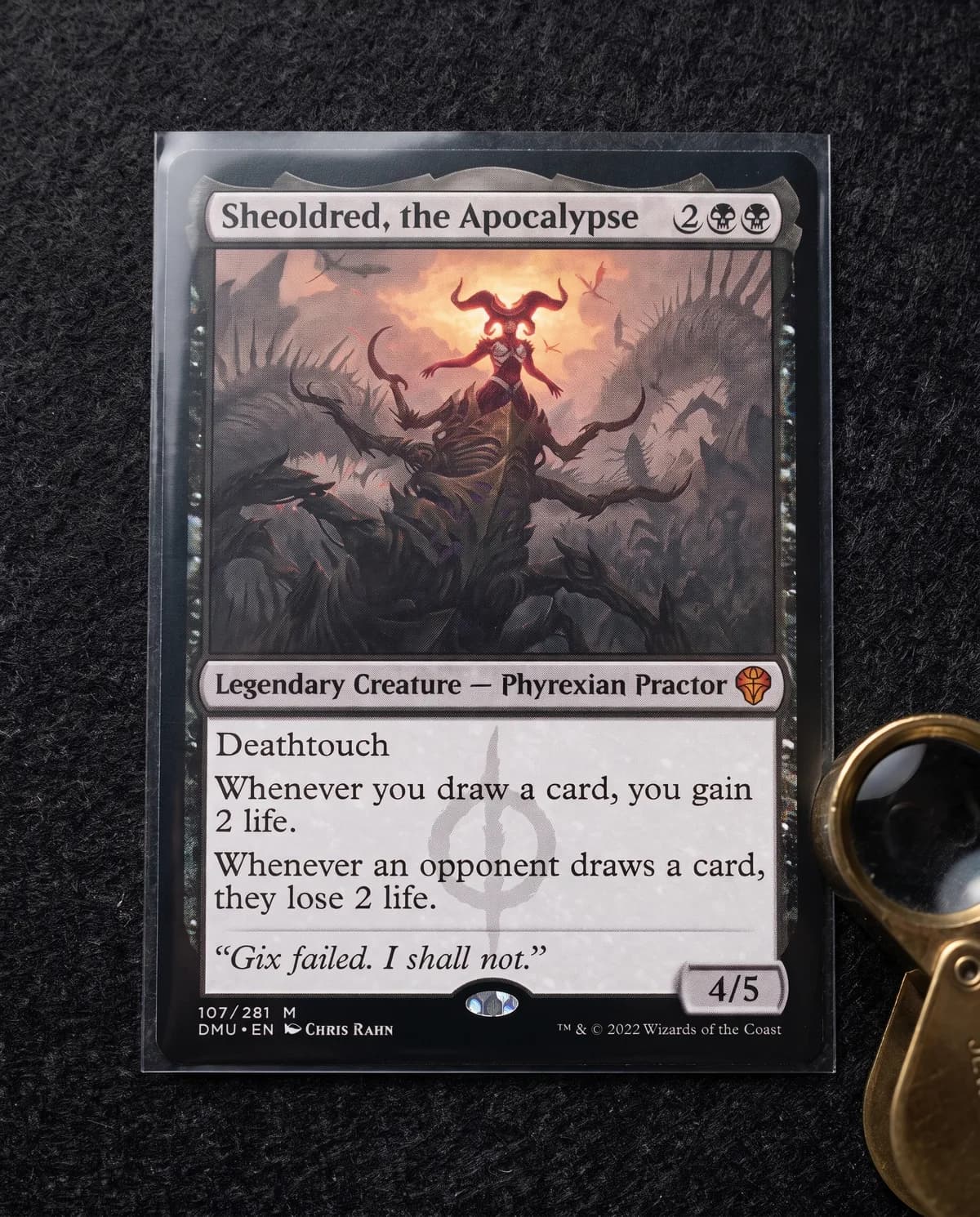 Sheoldred, the Apocalypse in a penny sleeve beside a jeweller's loupe, being checked for condition